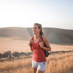 Smiling woman in casual attire enjoying a summer hike through golden fields at sunset.