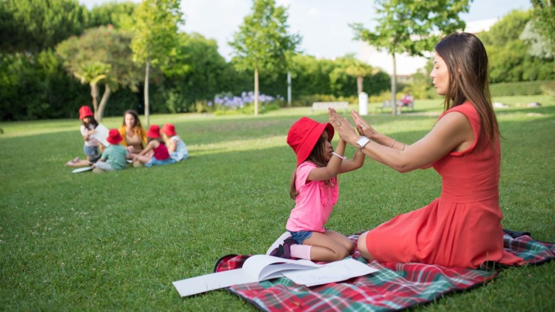 Joyful interaction between a child and woman in a sunny park, surrounded by nature and other children.
