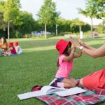 Joyful interaction between a child and woman in a sunny park, surrounded by nature and other children.